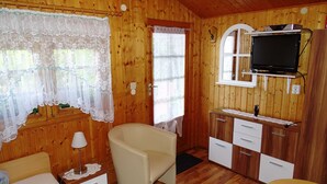 A cozy wooden room with a bed, a chair, a small table, a TV mounted on a cabinet, and a window with lace curtains.