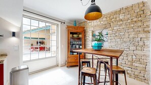 Un coin repas avec une table en bois, deux tabourets, un mur en pierre et, par une porte vitrée, une vue sur la cuisine.