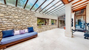 Un mur en pierre avec un banc en bois et des coussins, une porte en verre donnant sur une salle de sport et un sol en pierre.