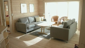 Ein gemütliches Wohnzimmer mit einem grauen Sofa, einem kleineren grauen Sessel und einem runden Couchtisch. An der Wand hängen zwei gerahmte Bilder, und durch die Glastür ist ein Korbstuhl zu sehen.