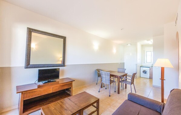A compact living space with a wooden entertainment unit, a sofa, a dining table with chairs, a washing machine, and a lamp. - Amazing apartment in Riumar-Deltebre with swimming pool (Riumar-Deltebre)