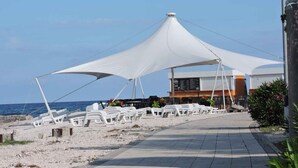 Ein Promenadenweg am Strand mit weißen Kunststoffstühlen und einer überdachten Sitzgelegenheit, der zu einem Gebäude mit einer Speisekarte führt.