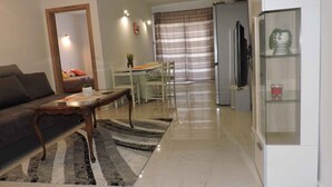 Ein Wohnzimmer mit einer Couch, einem Couchtisch und einem Essbereich mit Tisch und Stühlen. Im Hintergrund befinden sich ein Kühlschrank und ein Fernseher.