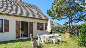 Une maison avec un toit en tuiles, des volets en bois et une façade blanche. On y trouve une terrasse avec une table et des chaises, et un arbre en arrière-plan.