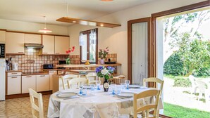 Une cuisine bien éclairée, dotée d’appareils électroménagers modernes, un coin repas avec une table dressée pour quatre personnes, et une grande fenêtre offrant une vue sur le jardin.