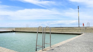 Une piscine en béton avec des échelles en métal, un chemin pavé et une personne qui marche au loin.