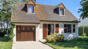 Une maison à deux étages, avec un toit recouvert de tuiles, une porte de garage en bois et un petit jardin agrémenté de fleurs jaunes.