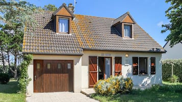 A two-story house with a tiled roof, a wooden garage door, and a small garden with yellow flowers.
