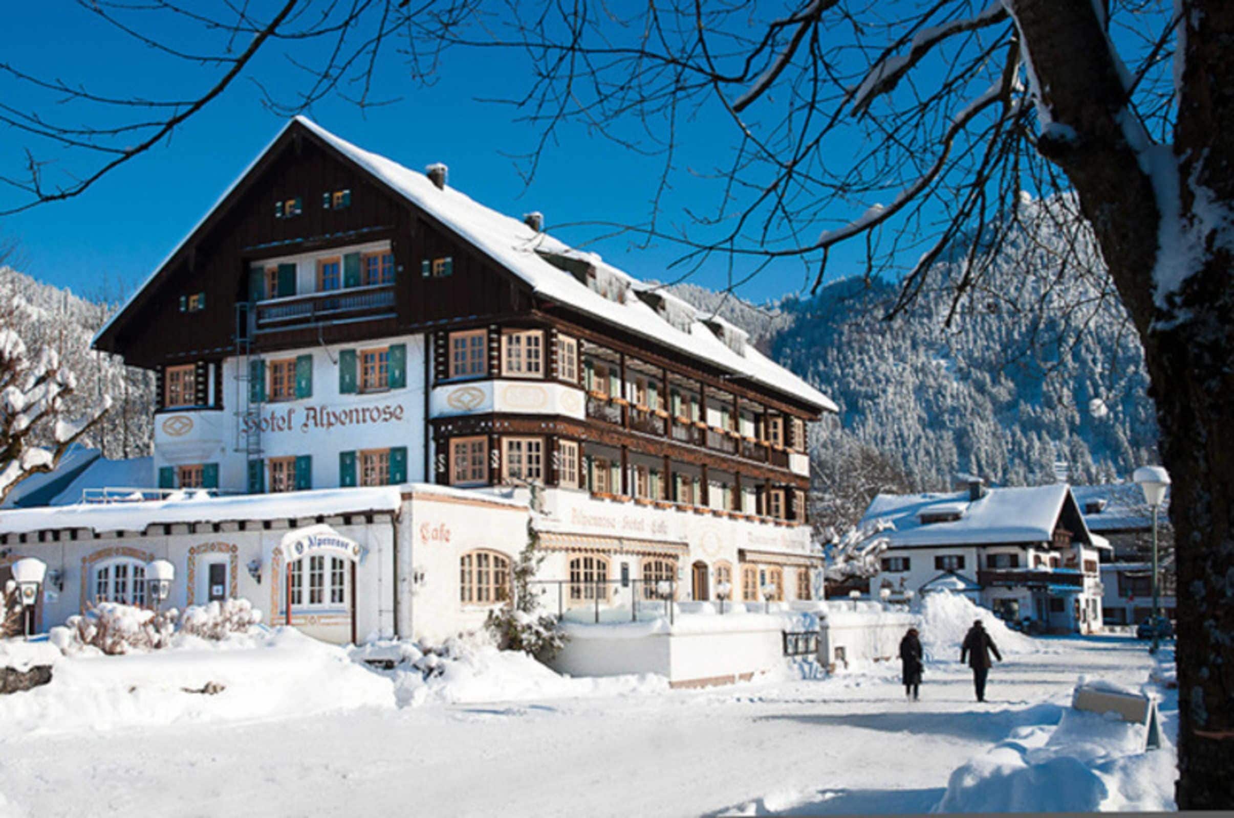 A traditional wooden building with multiple windows and balconies, a sign indicating it's a hotel, and a snowy landscape with mountains in the background.
