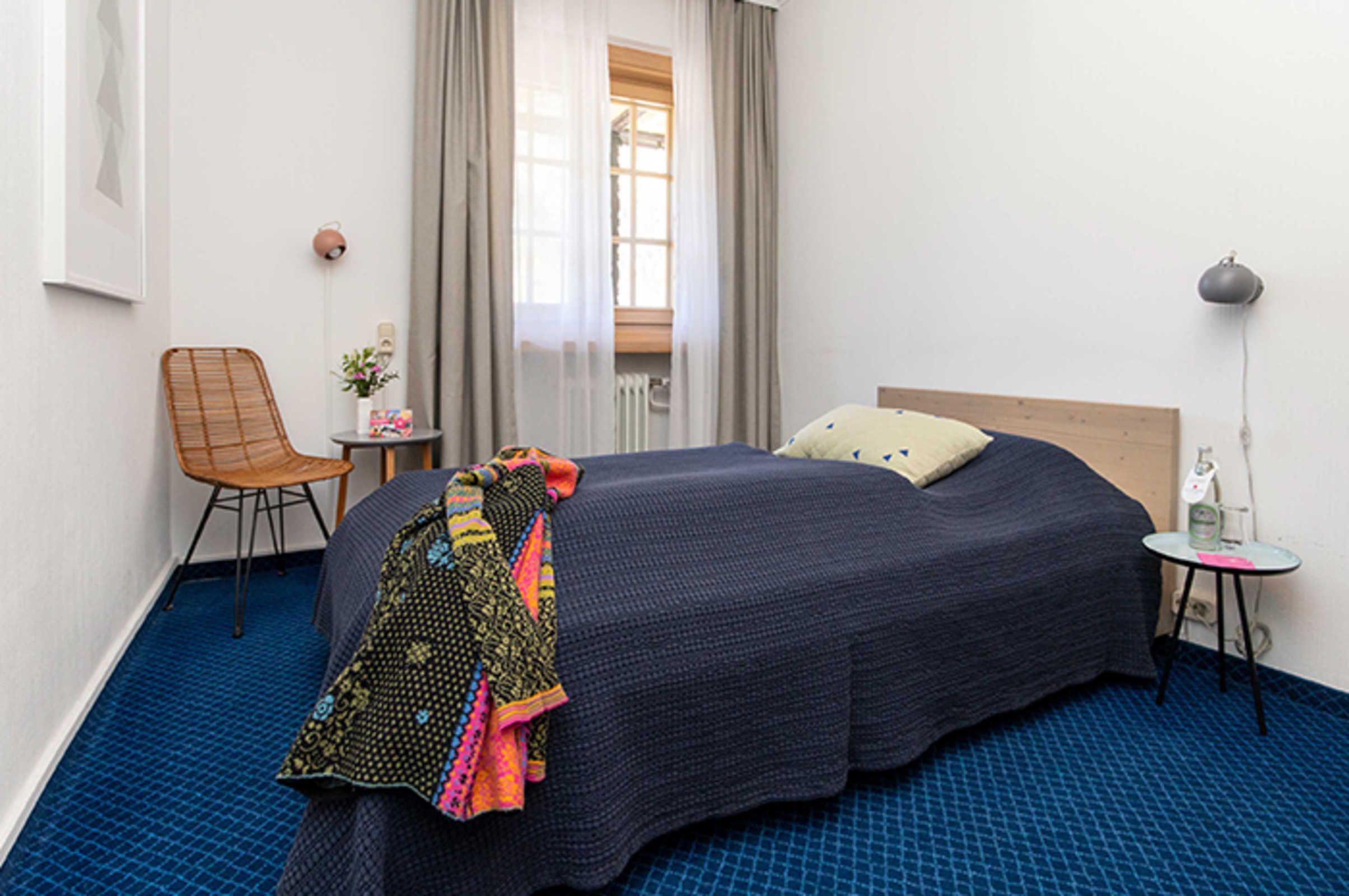 A hotel room with a bed, a chair, a small table, a lamp, and a window with curtains.