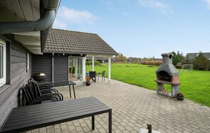 A patio area with a table and chairs, a stone oven, and a view of a green lawn and houses in the background. - Awesome home in Idestrup with sauna (Idestrup)