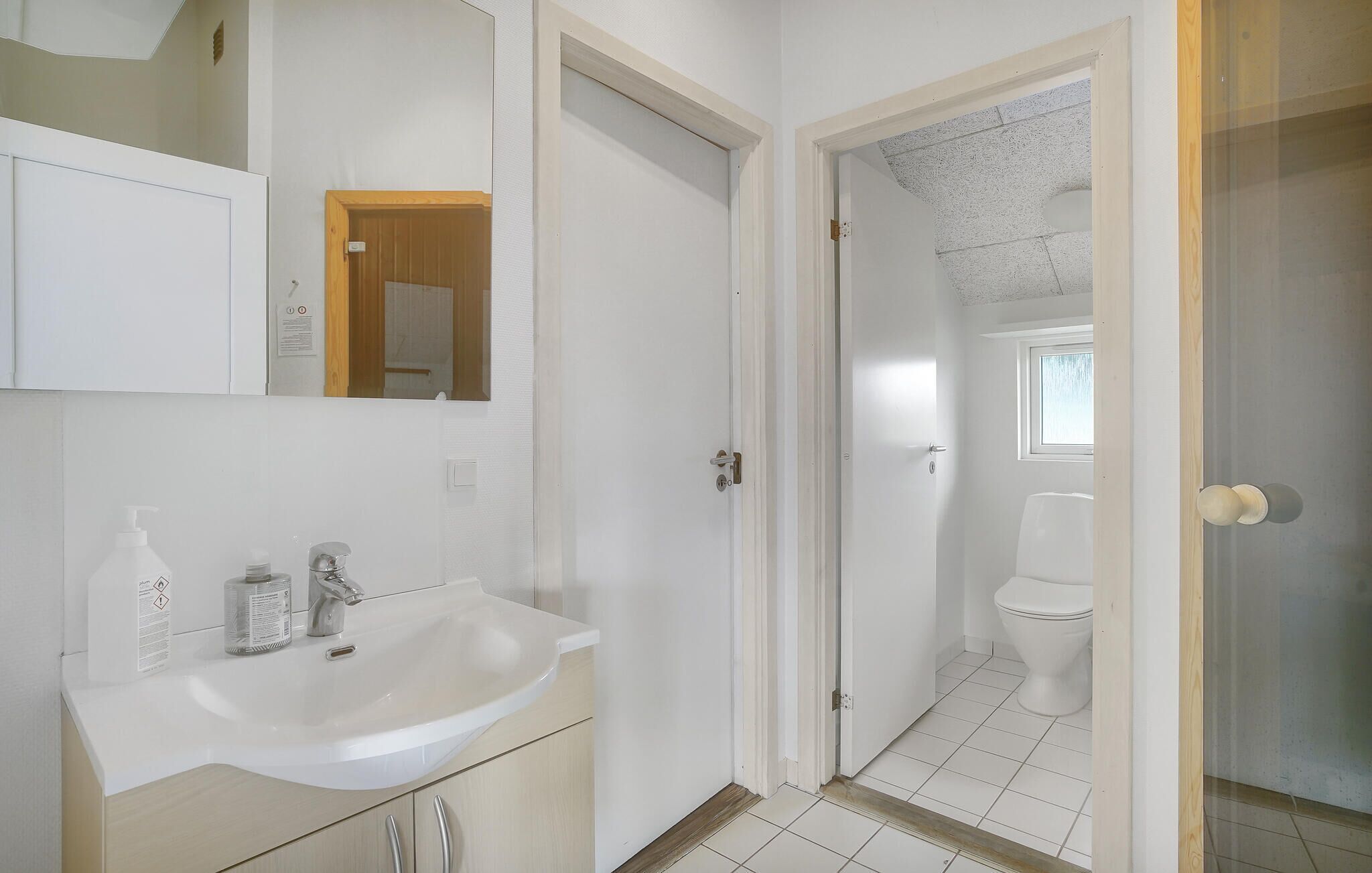 Un bagno con lavandino, specchio e mobiletti bianchi. Si apre una porta verso il bagno e la zona doccia.