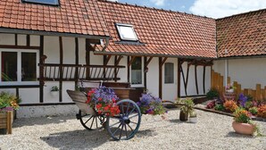 En traditionel bindingsværkshus med en grusbelagt gårdsplads, en trævogn med blomster og et rødt tegltag.