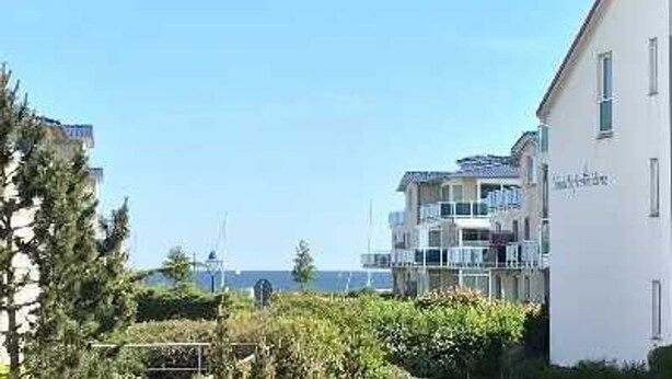 A coastal residential area with modern buildings, including a two-story house with a balcony and a white building with a sign. - (164) 2-room apartment - apartment complex Yachthafenresidenz (Kühlungsborn)