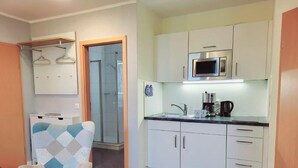 A compact kitchen with white cabinets, a microwave, and a coffee maker. There's a chair with a geometric pattern and a door leading to another room. - (164) 2-room apartment - apartment complex Yachthafenresidenz (Kühlungsborn)