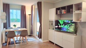 A modern living room with a flat-screen TV, a dining table with chairs, and a bookshelf. - (164) 2-room apartment - apartment complex Yachthafenresidenz (Kühlungsborn)