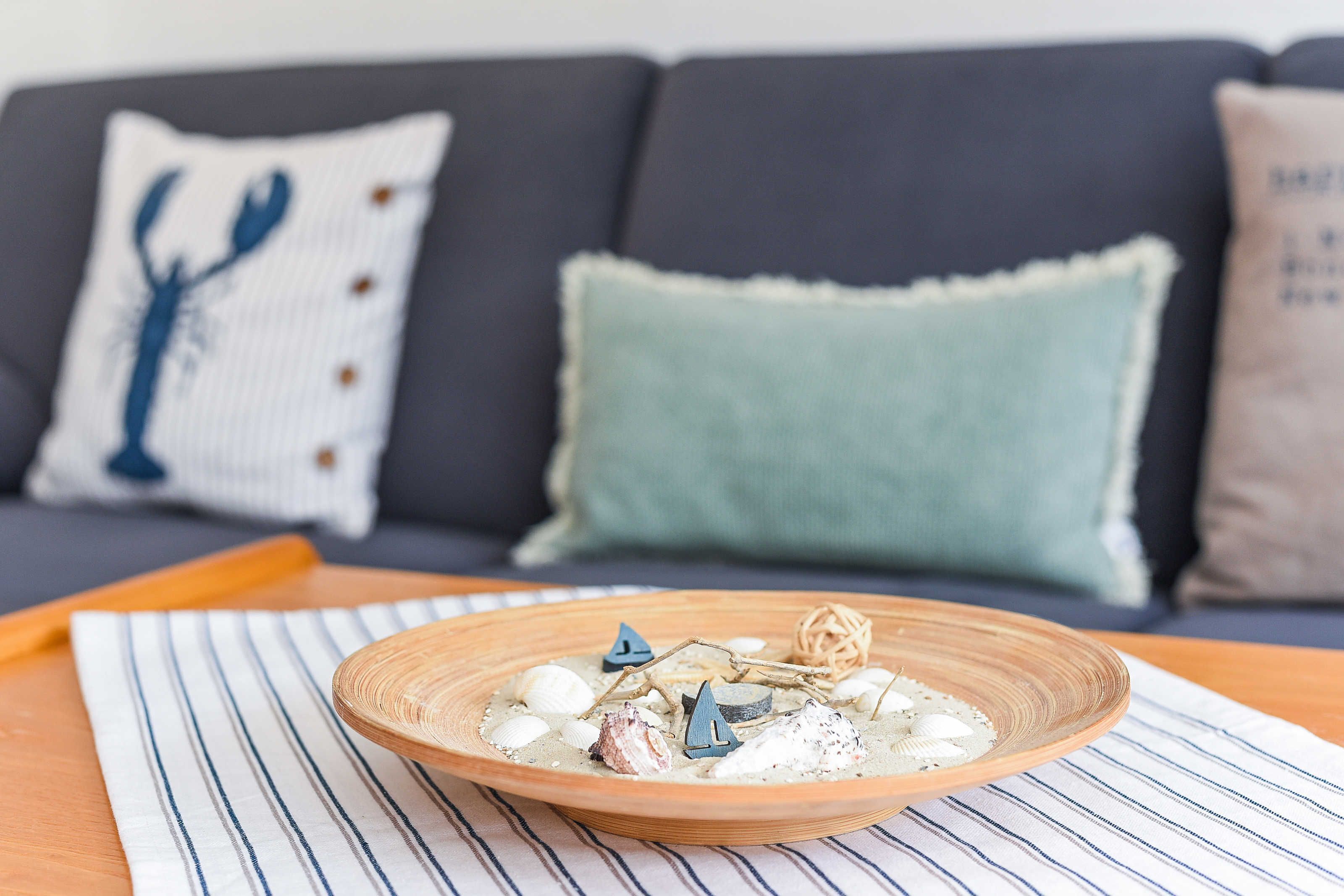 Un bol en bois contenant des objets décoratifs, posé sur un tapis à rayures, sur une table devant un canapé sombre agrémenté de coussins à motifs.