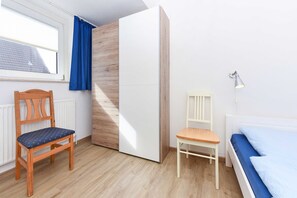 Une chambre avec un plancher en bois, un lit avec un matelas bleu, une armoire blanche, une chaise en bois avec un coussin bleu et une fenêtre avec des rideaux bleus.