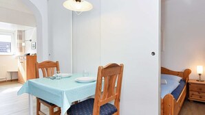 Een eethoek met een tafel voor twee personen, houten stoelen, een hanglamp en een schuifdeur die leidt naar een slaapkamer met een bed en nachtkastje.