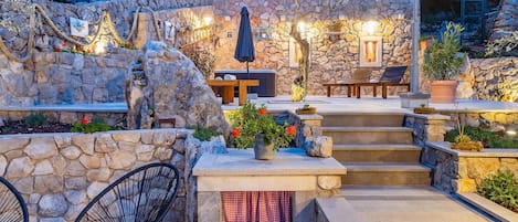 A stone-walled outdoor patio with steps leading to a seating area, featuring a table and chairs, and potted plants.