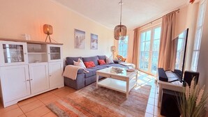 A living room with a sofa, coffee table, and a TV. There is a cabinet with glass doors and a hanging lamp. - (5) 3-room apartment south-facing balcony - apartment house Atlantic (Kühlungsborn)