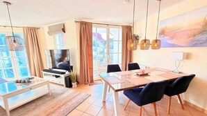 A modern living room with a TV, dining table, chairs, and a large window. - (5) 3-room apartment south-facing balcony - apartment house Atlantic (Kühlungsborn)