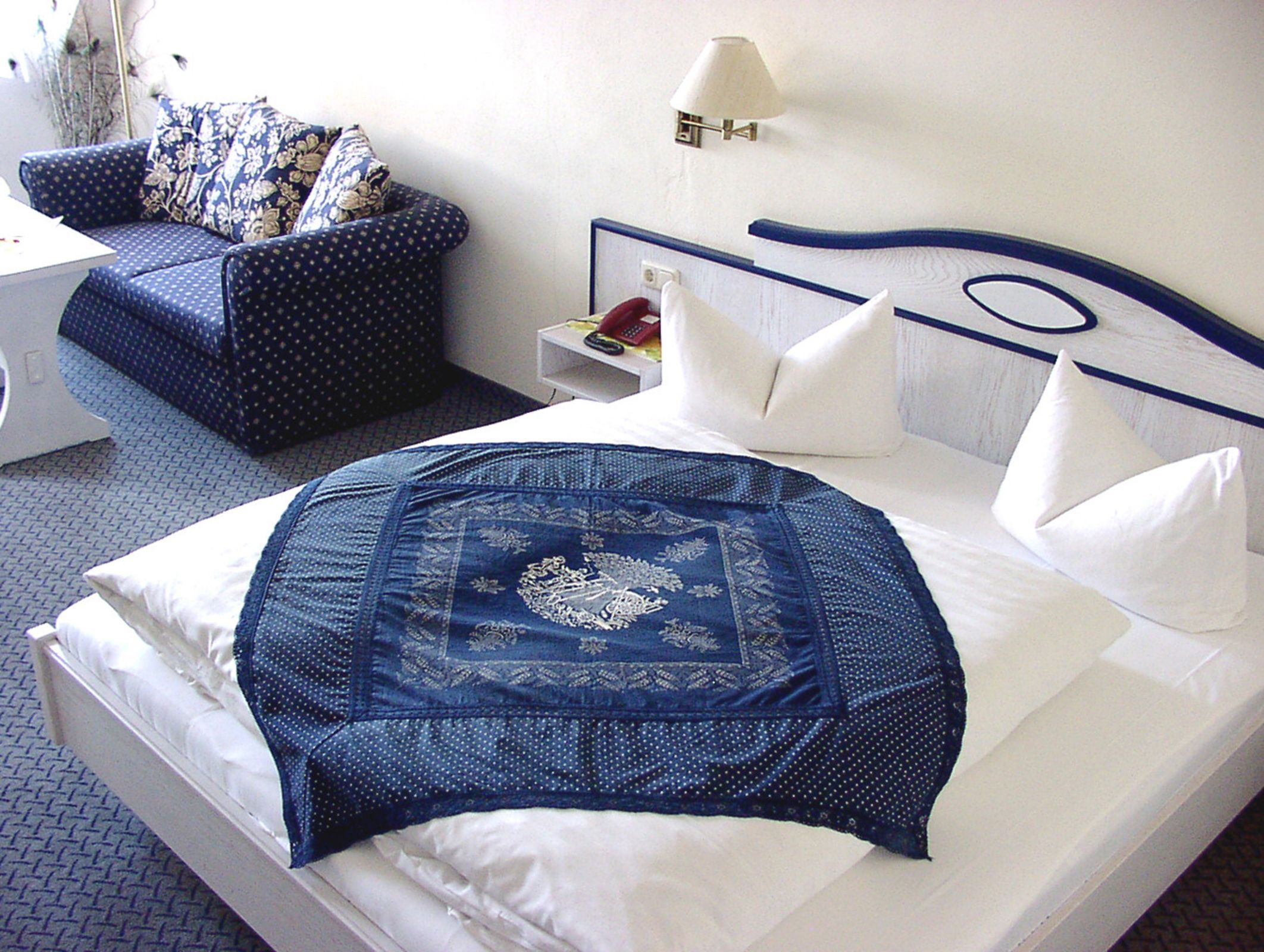Una cama bien hecha con una colcha azul y blanca estampada, un almohadón blanco y otro a juego en la cabecera. Se ve una pequeña mesita de noche con una lámpara y un libro. En el fondo, un sofá azul con cojines florales.
