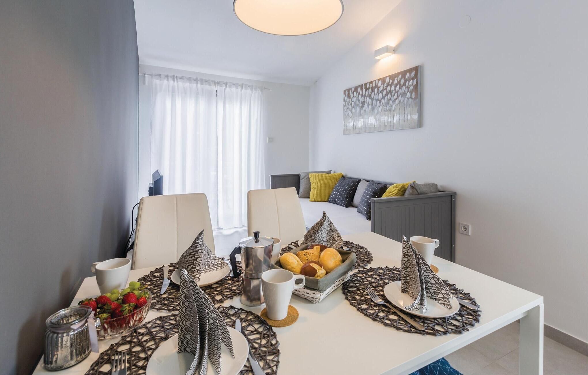 A modern hotel room with a dining area featuring a white table set with cutlery, plates, and a fruit bowl. A bed with a gray headboard and colorful pillows is visible in the background.