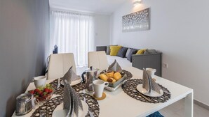 Et moderne hotelværelse med spiseplads, hvor et hvidt bord er dækket med bestik, tallerkener og en skål med frugt. En seng med gråt sengegavl og farverige puder ses i baggrunden.