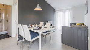 Et moderne spiseområde med et hvidt bord dækket til fire personer, en grå kommode med en seng og et bruseområde.