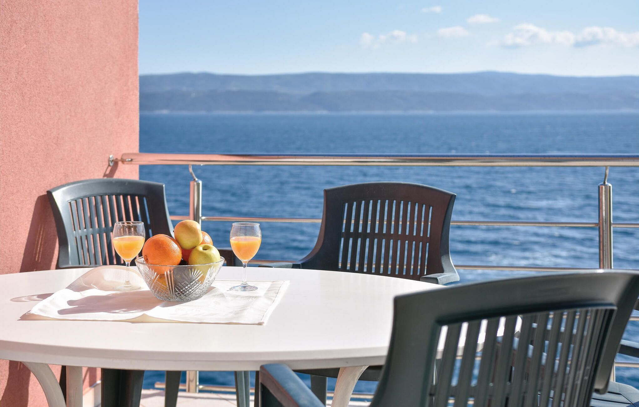 Een ronde tafel met een witte tafeldoek, twee zwarte stoelen, een schaal met fruit en twee glazen sinaasappelsap op een balkon met uitzicht op zee.