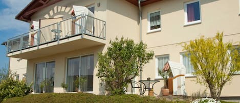 A two-story building with a balcony, large windows, and a garden with various plants and flowers.