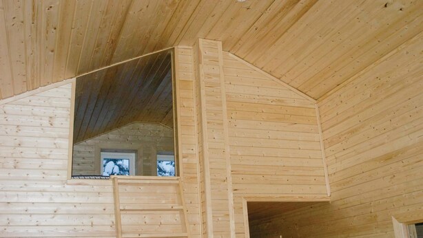 Ein Holzhütten-Innenraum mit Schlafzimmer im oberen Bereich, einer Leiter zum Schlafbereich und einem Fenster mit Blick auf schneebedeckte Bäume.
