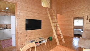 Innenansicht einer Holzhütte mit Küche, einem an der Wand montierten Fernseher, einer Holzleiter, einem kleinen Tisch mit Zeitschriften und einem Sessel.