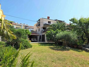 Un quartiere residenziale con una casa a due piani, dotata di un balcone, un piccolo patio e un prato curato con arbusti e alberi di diverse varietà.