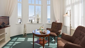 A room with a large window, a television, a small table with a decorative item, a brown armchair, and a floor lamp. - Double room comfort with sea view - Strandvilla Imperator | Hotel & (Heringsdorf)