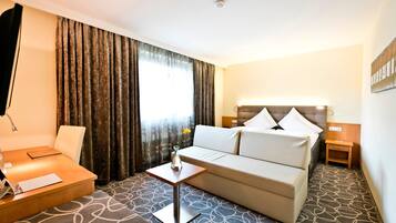 Quarto de hotel com cama, sofá, uma mesinha com um vaso, luminária e escrivaninha com teclado.