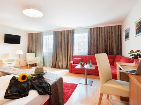 Une chambre d’hôtel moderne avec un lit, un canapé rouge, un bureau avec un ordinateur portable, un téléviseur fixé au mur et une grande fenêtre avec des rideaux.