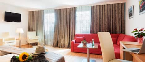 A modern hotel room with a bed, a red sofa, a desk with a laptop, a TV mounted on the wall, and a large window with curtains.