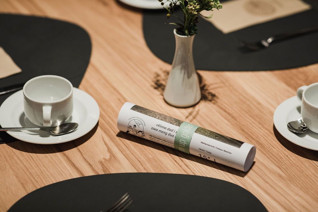 Ein Tisch mit einer weißen Tasse, einem Löffel, einer gerollten Speisekarte und einer Vase mit Blumen.