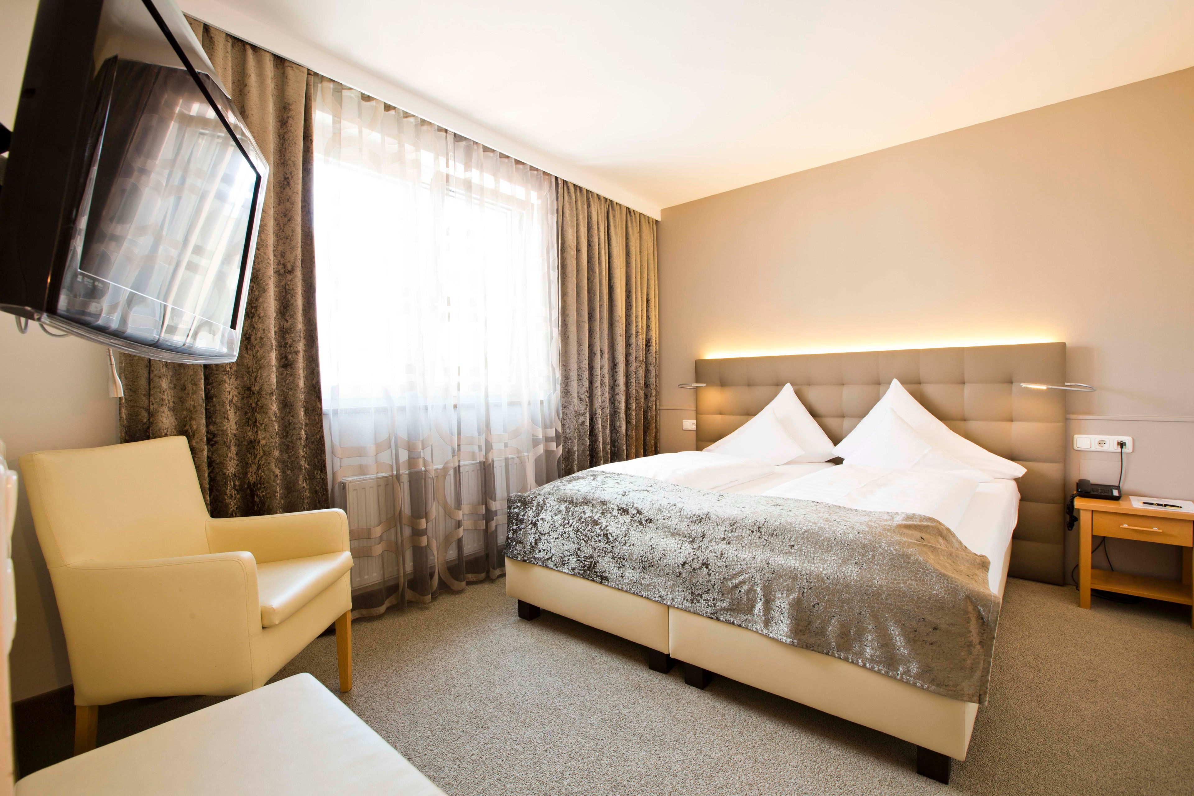 Habitación de hotel con cama, una silla, mesita de noche, televisor y ventana con cortinas.