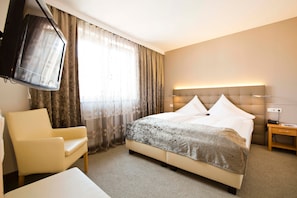 Ett hotellrum med en säng, en stol, ett sängbord, en tv och ett fönster med gardiner.