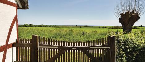 A wooden gate with a metal latch, a white building with red accents, and a field with a tree in the background.
