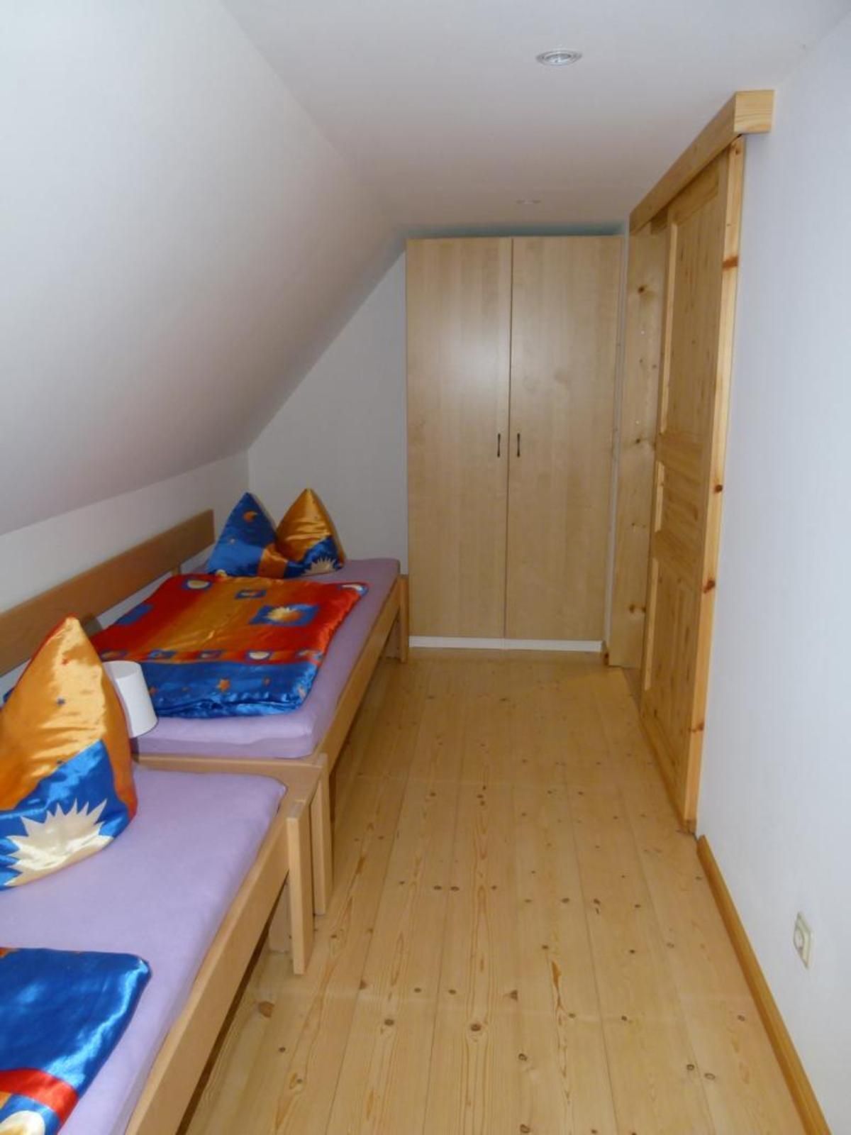 A small, narrow room with a wooden floor, a bed with colorful bedding, and a wooden wardrobe.