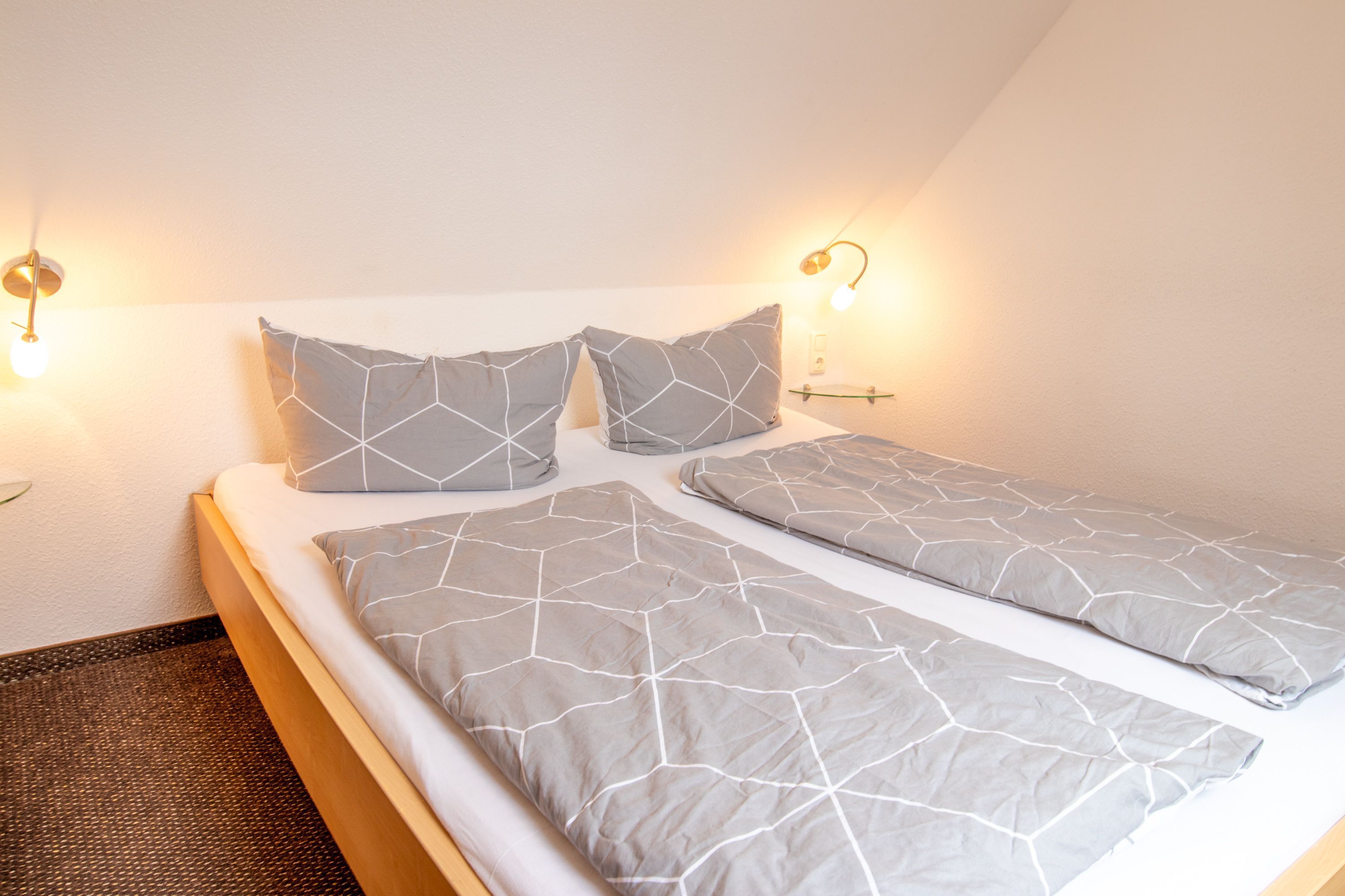 A neatly made bed with a geometric-patterned bedspread and pillows, two wall-mounted reading lamps, and a small bedside table.