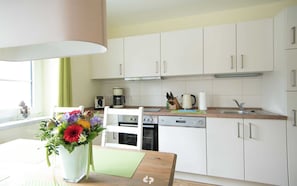 Eine moderne Küche mit weißen Schränken, einem Esstisch mit Blumenvase und einem Fenster mit Vorhängen.