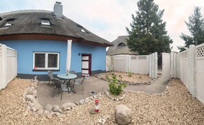 Ein Haus mit einem Strohdach, blauer Fassade, einer kleinen Terrasse mit Tisch und Stühlen sowie einem weißen Zaun, der das Grundstück umgibt.