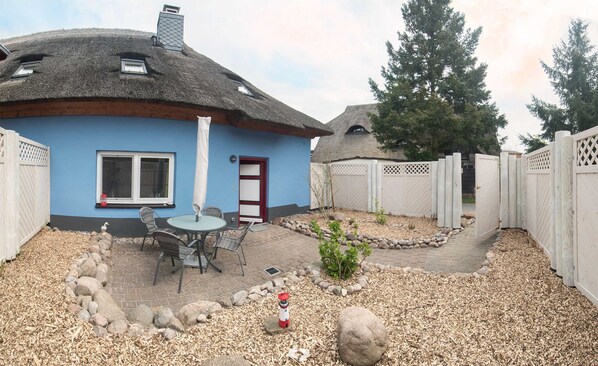 A house with a thatched roof, a blue exterior, a small patio with a table and chairs, and a white fence surrounding the property.