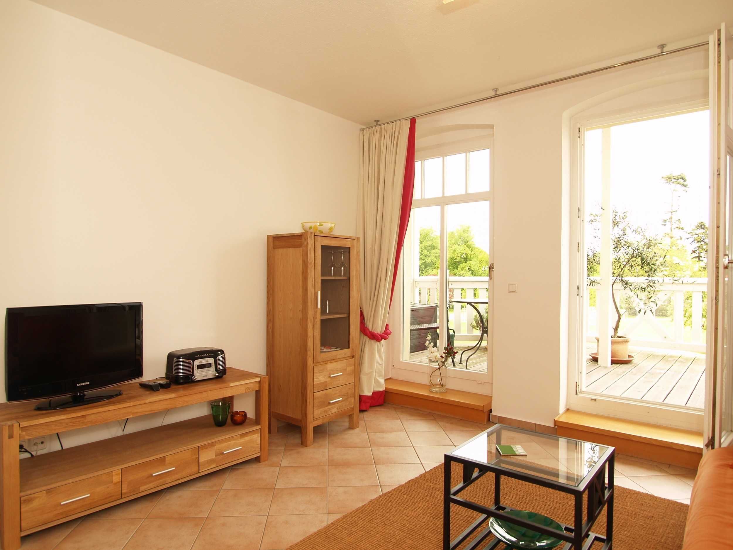 Ein Wohnzimmer mit Fernseher, Radio, Holzschrank, Glastisch und Blick ins Freie durch ein großes Fenster.