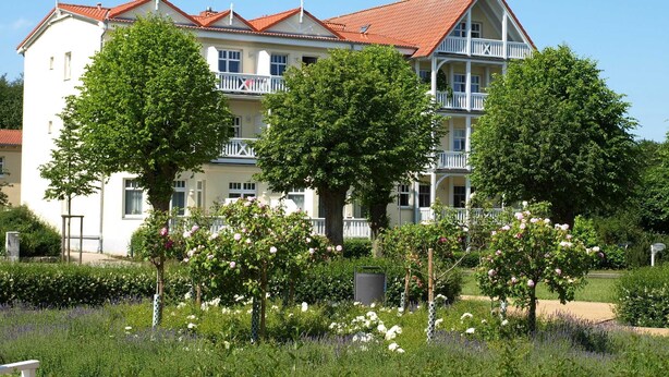 En bygning med flere etager, rødt tegltag, hvide balkoner og frodig vegetation i forgrunden.